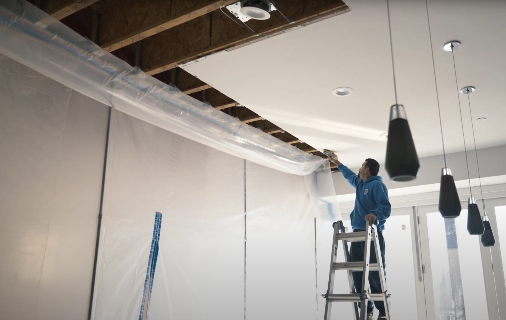 Water damage restoration professional on ladder working on ceiling repair with protective plastic sheeting
